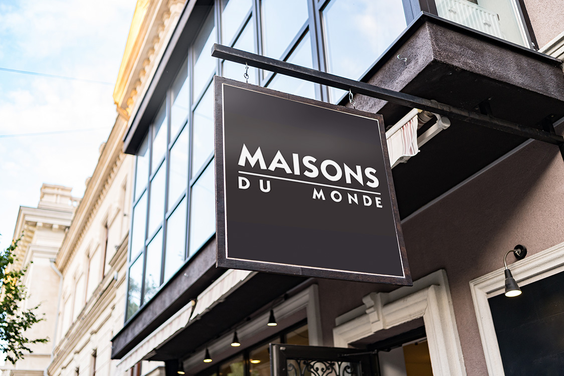 Un cartel que cuelga de una pared en plena calle, donde una tipografia blanca resalta por encima de un fondo oscuro, en esta se puede leer Maisons du Monde