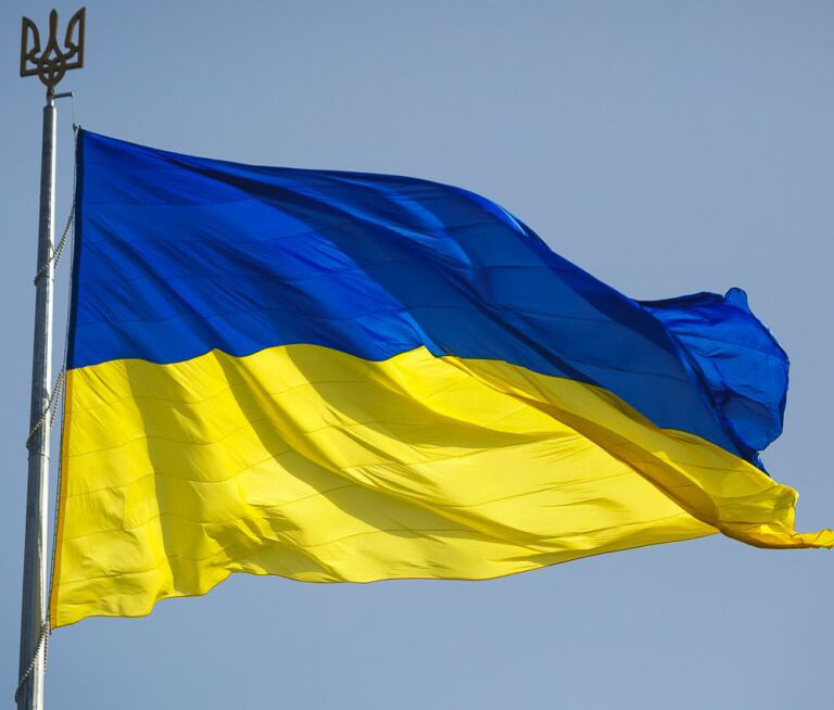 Imagen con bandera ucraniana, hondea en el cielo mientras esta atada al poste, este en en la punta tiene el tridente en representación al escudo de ucrania.